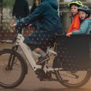 Adult riding a white cargo bike with two helmeted children seated in the front box on a city street. A dotted graphic overlay and white bicycle icons appear across the image, with a warm light flare in the lower right corner.