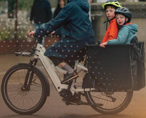 Adult riding a white cargo bike with two helmeted children seated in the front box on a city street. A dotted graphic overlay and white bicycle icons appear across the image, with a warm light flare in the lower right corner.