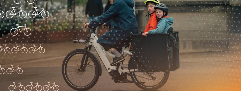 Adult riding a white cargo bike with two helmeted children seated in the front box on a city street. A dotted graphic overlay and white bicycle icons appear across the image, with a warm light flare in the lower right corner.