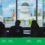 Advocates sit in silhouette inside the California State Capitol, facing a bright window with a view of Sacramento’s Capitol dome and downtown buildings. A green graphic band with white bicycle icons runs along the bottom of the image.