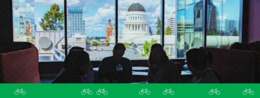 Advocates sit in silhouette inside the California State Capitol, facing a bright window with a view of Sacramento’s Capitol dome and downtown buildings. A green graphic band with white bicycle icons runs along the bottom of the image.