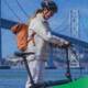 A cyclist with a backpack walks alongside an e-bike on San Francisco’s waterfront, with the Bay Bridge in the background, overlaid with blue halftone texture and green graphic waves.